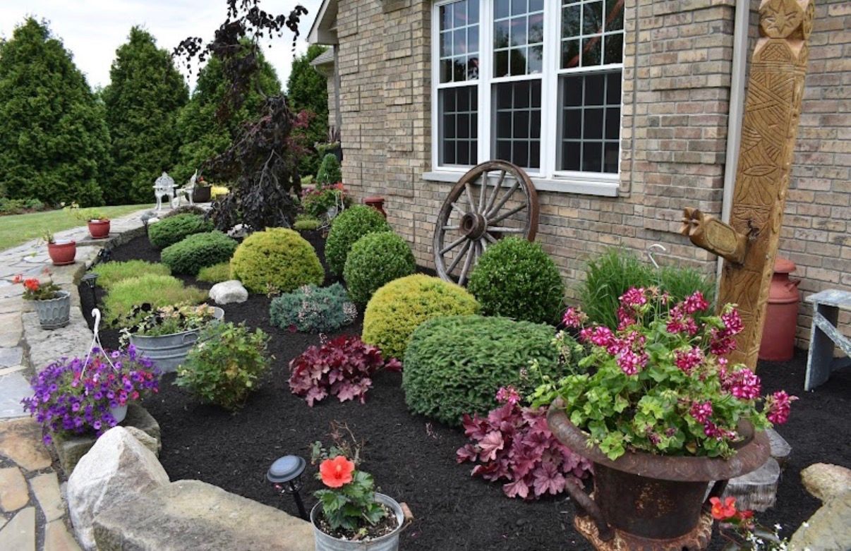 A landscaped garden bed with colorful flowers, rounded shrubs, a wooden wagon wheel, and carved wood sculpture near a house.