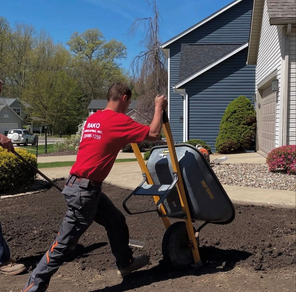 A Bako Landscaping worker in a red shirt and gray work pants uses a wheelbarrow to spread mulch in a residential yard.