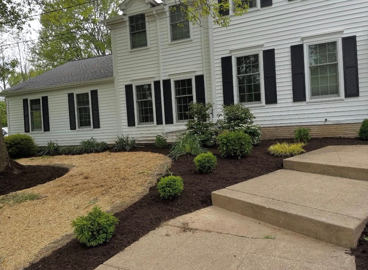 A white house exterior with black shutters, a concrete walkway, mulch, gravel, and small landscaping shrubs.