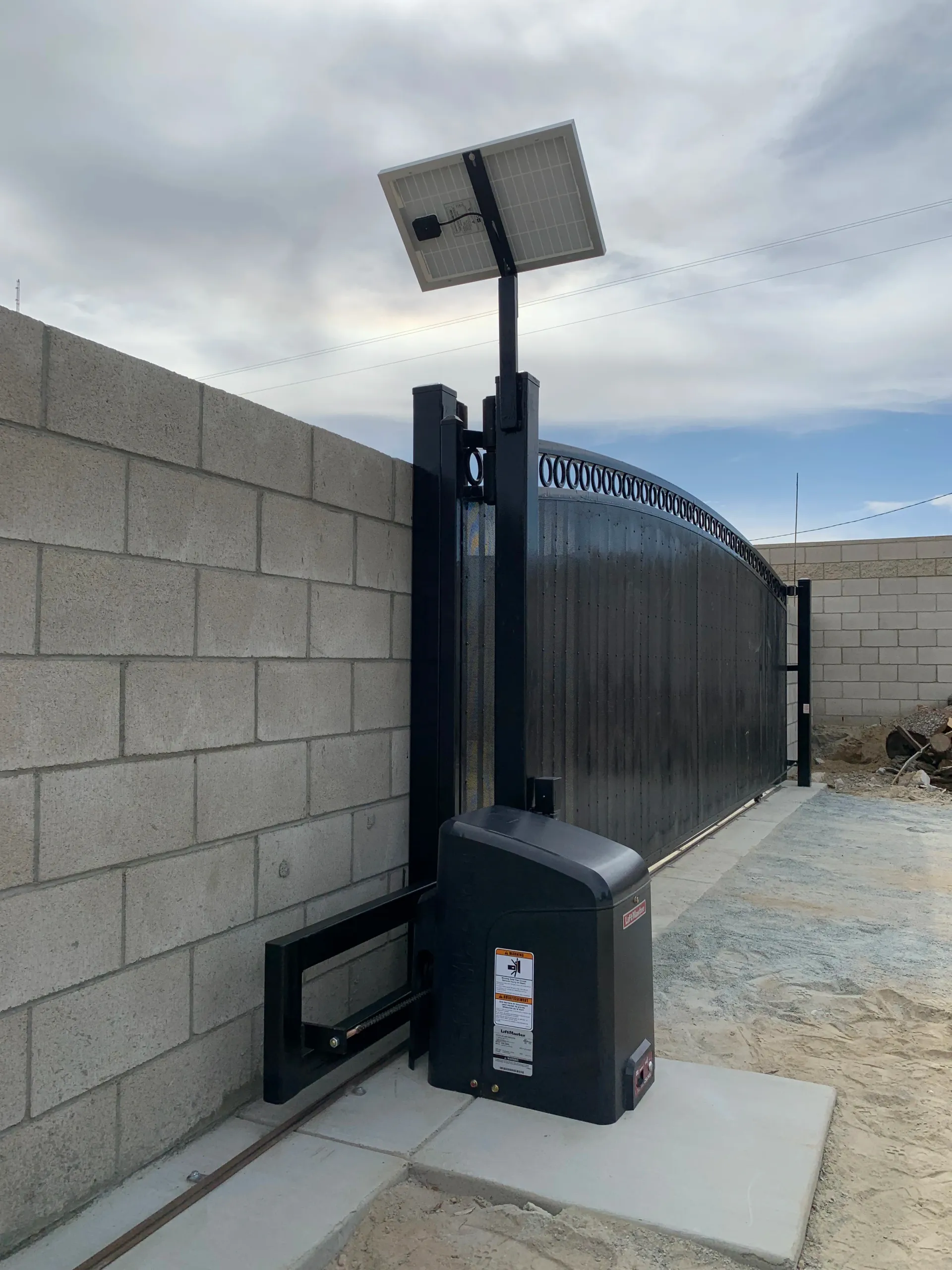 Solar-powered gate opener beside a black sliding gate and cinderblock wall.