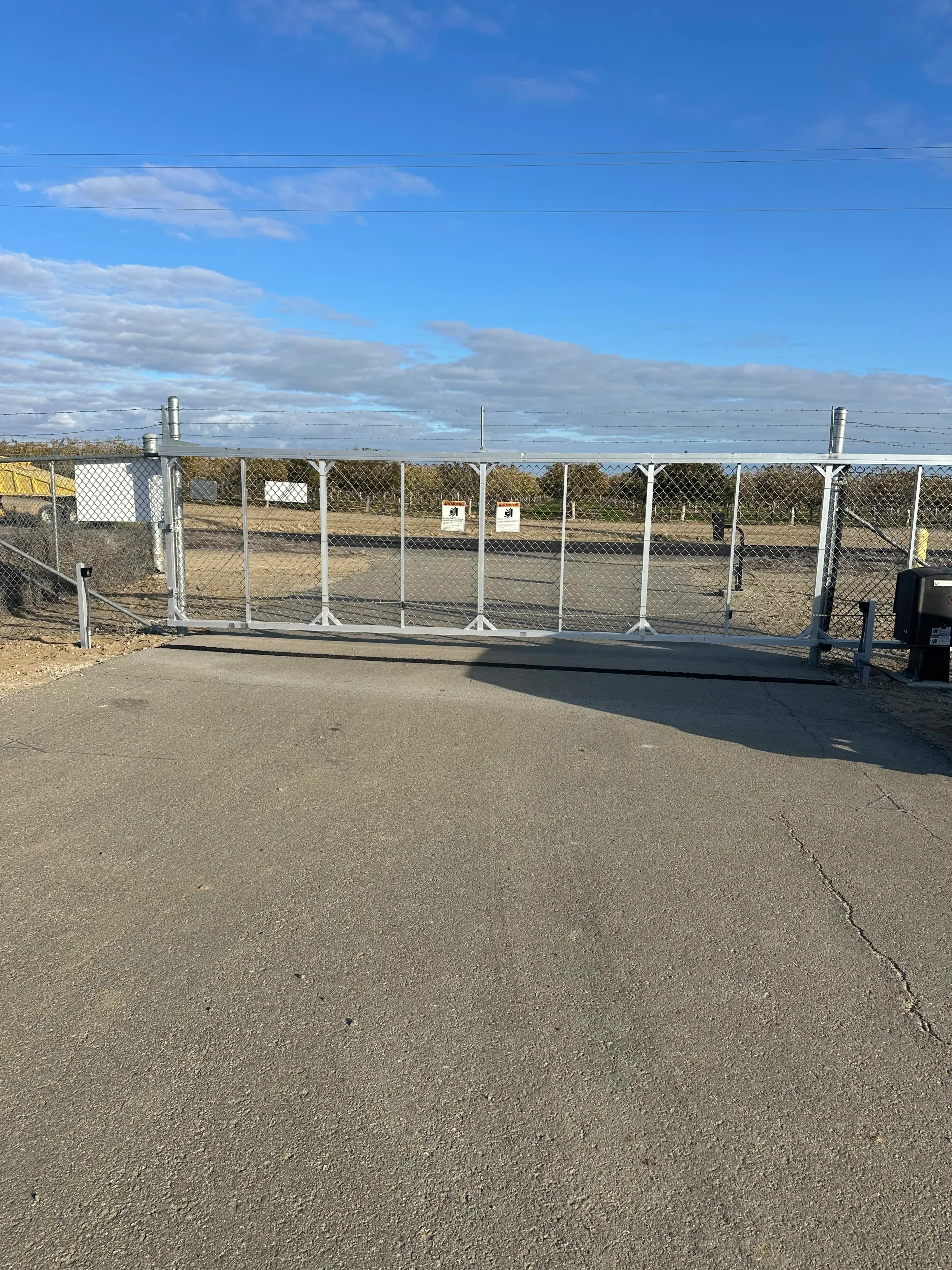 Chain-link gate blocking a paved road under a blue sky with scattered clouds in Kern County, CA