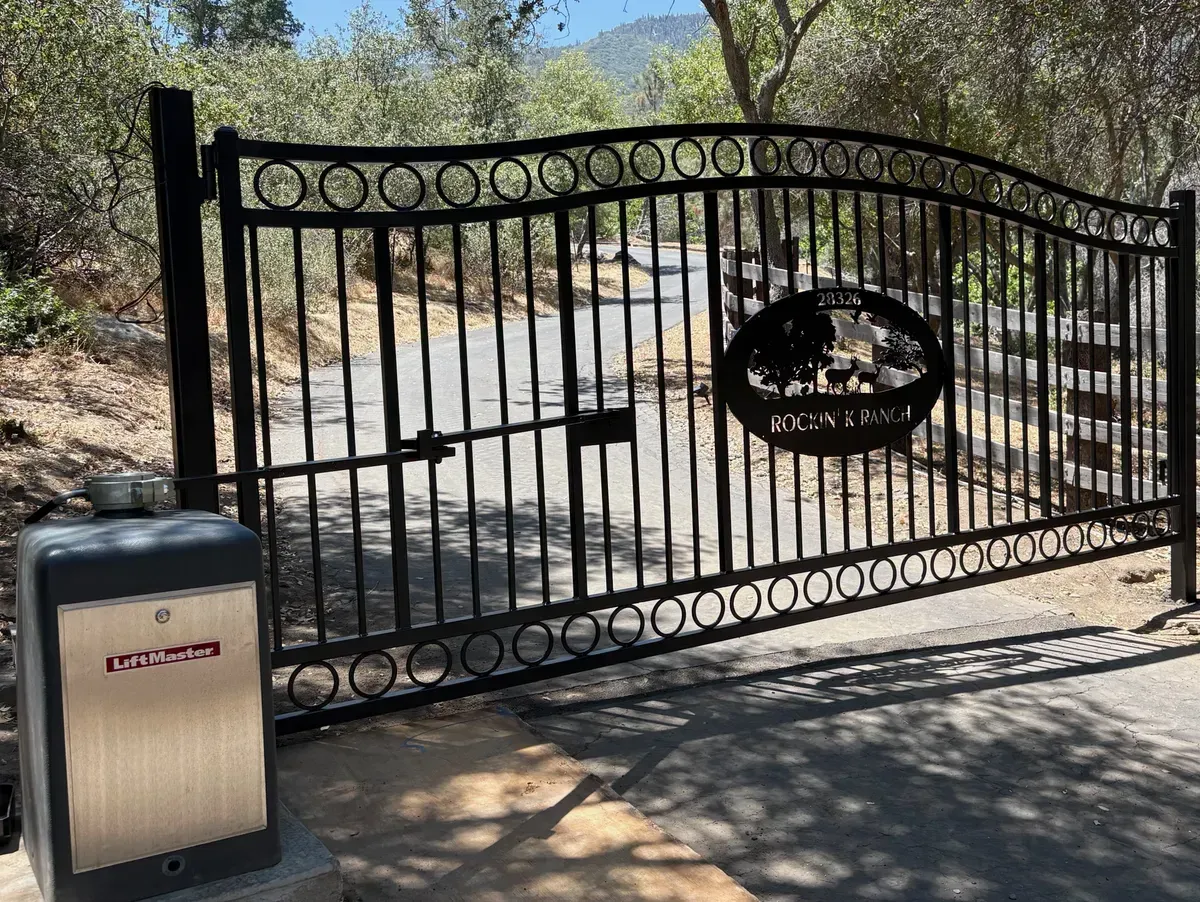 Black wrought-iron driveway gate with a stone pillar and gate opener, set by a road in a sunny rural area