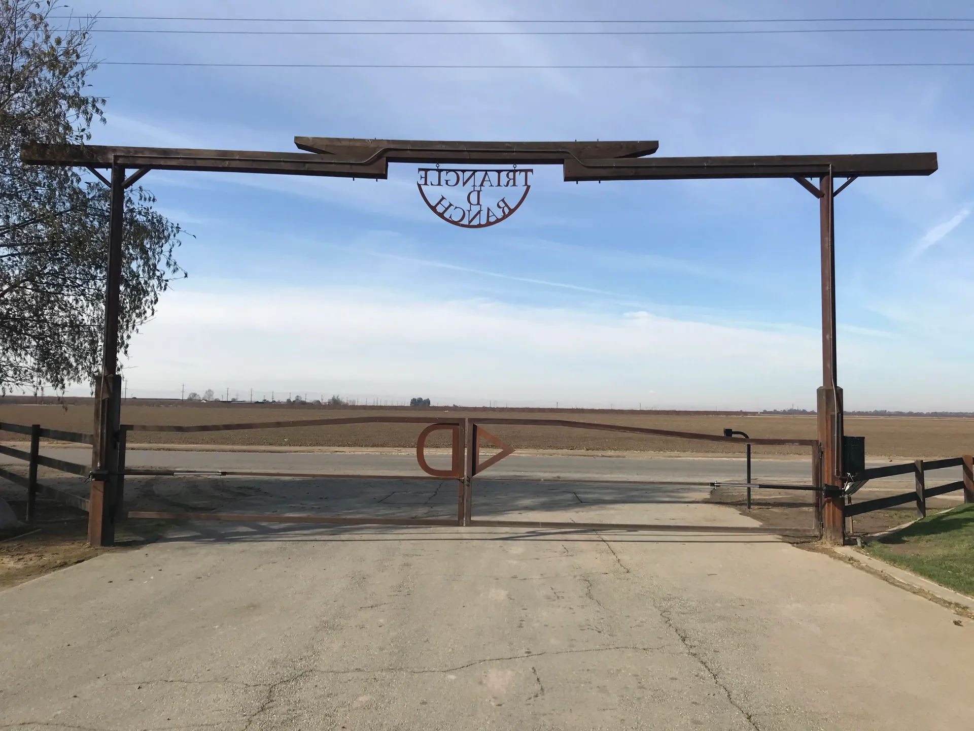 Rural entrance gate with wooden arch and closed fence across a dirt road under a clear blue sky