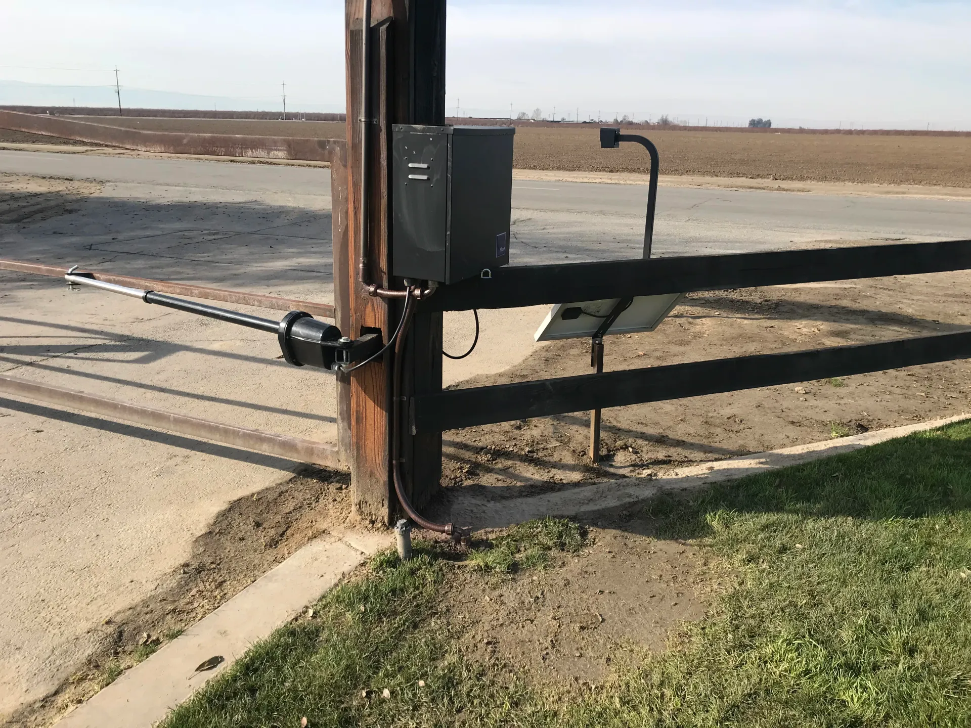 Black metal farm gate with electric opener beside a dirt road in a flat rural field