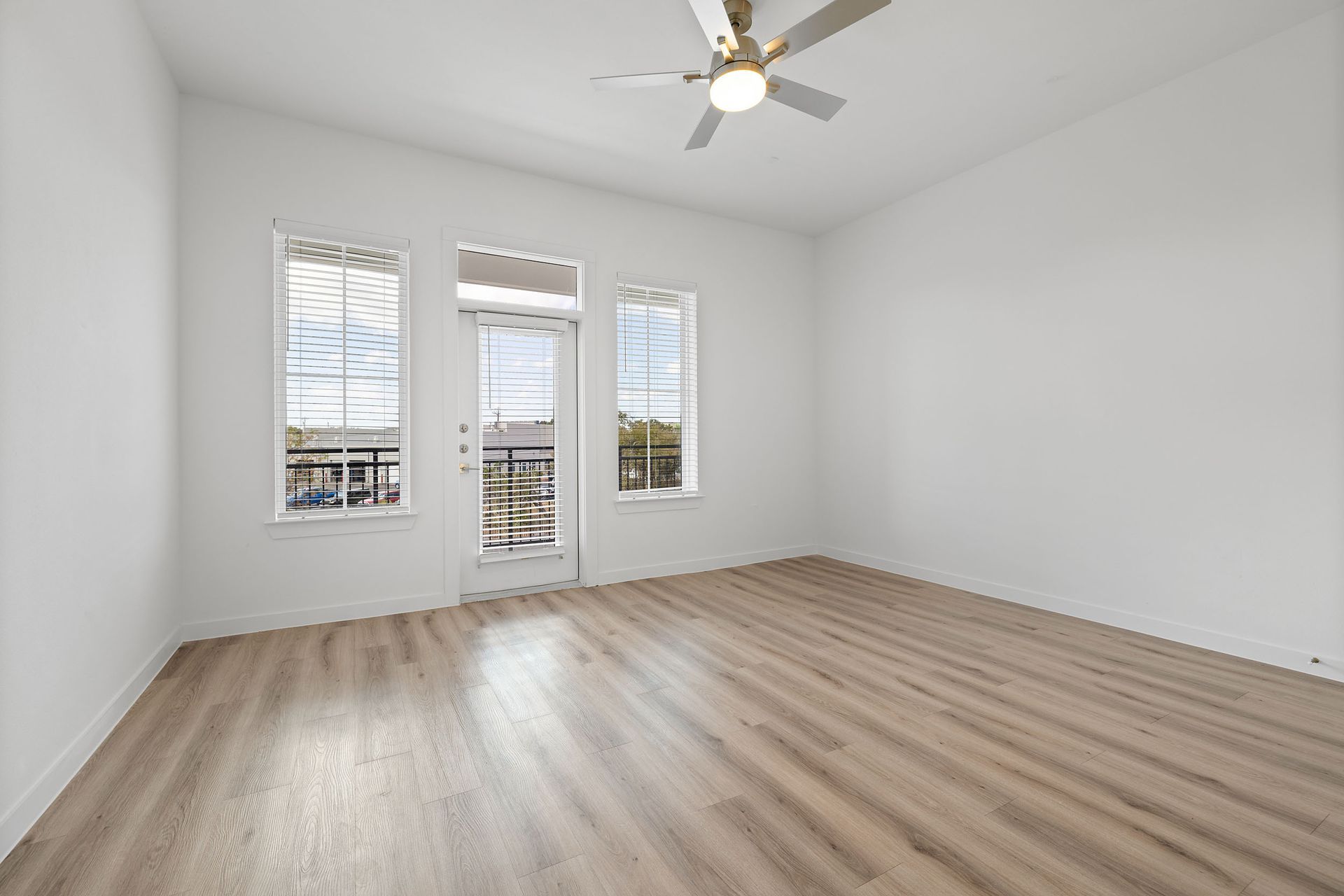 An empty living room with hardwood floors, 2 windows, balcony door, and a ceiling fan at The Everett, offering apartments for rent in Northwest Austin, TX.