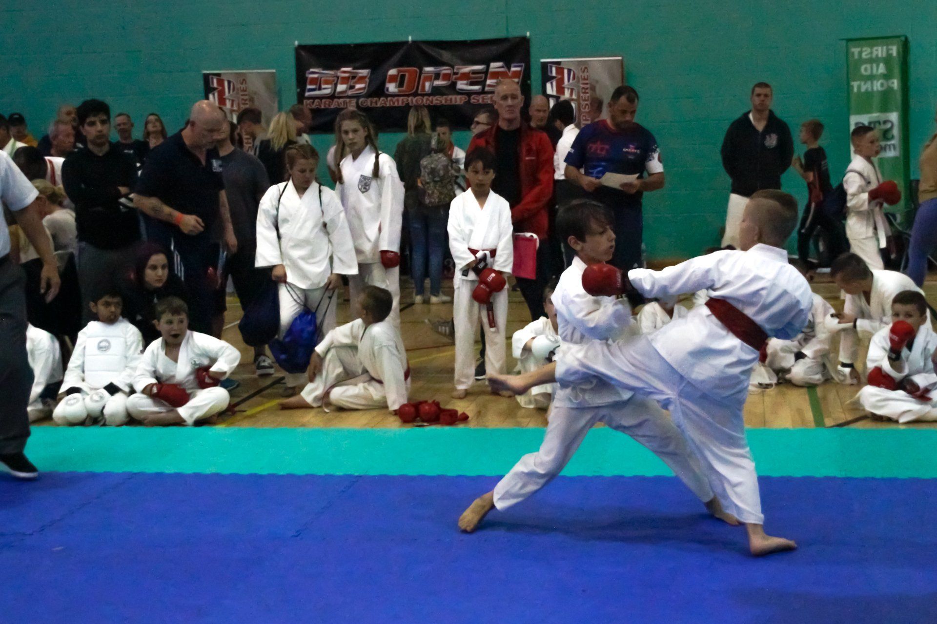 Two martial artists spar on blue mats while spectators watch in an indoor gymnasium.