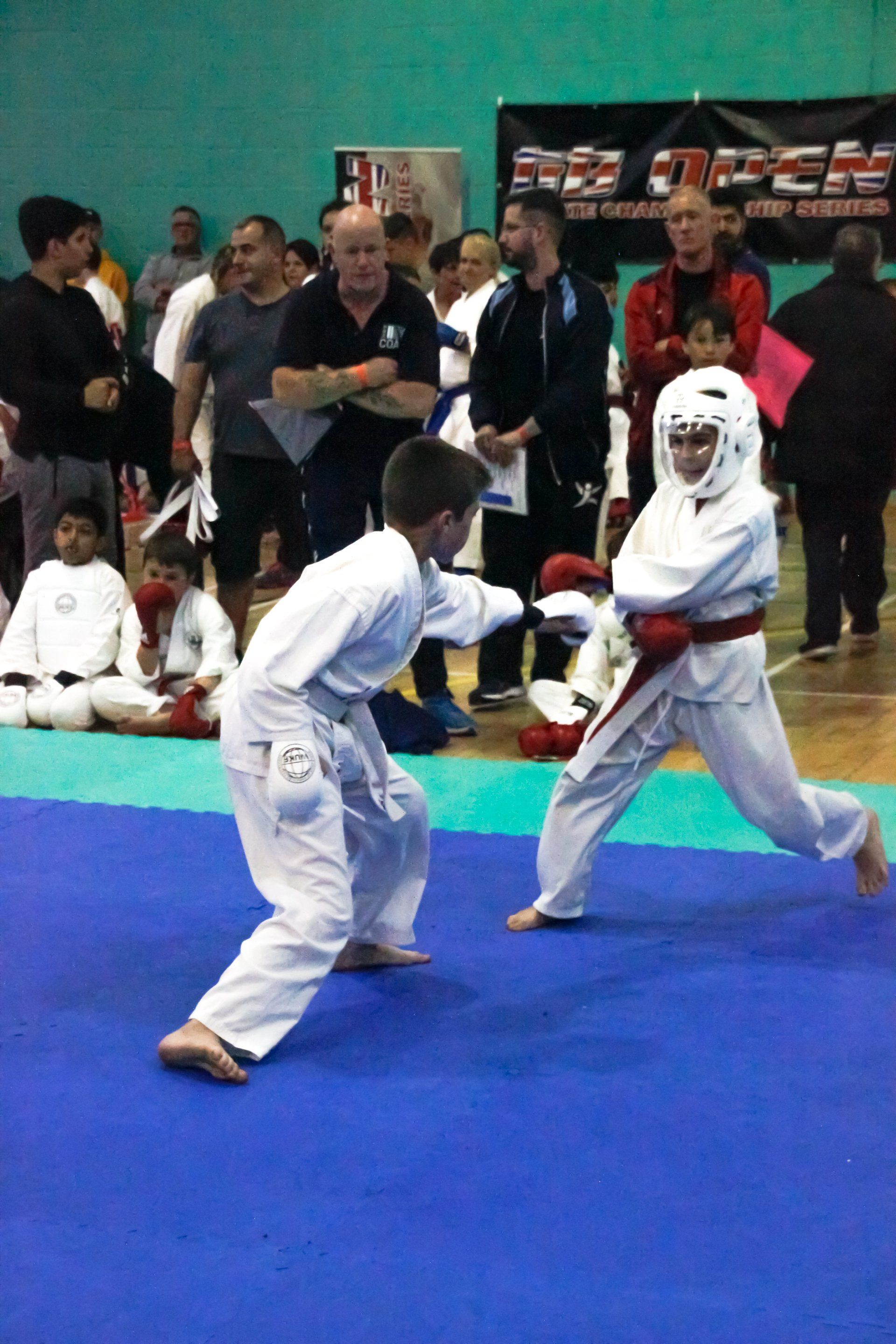 Two children in white martial arts uniforms spar on a blue mat in a gym, with spectators watching.