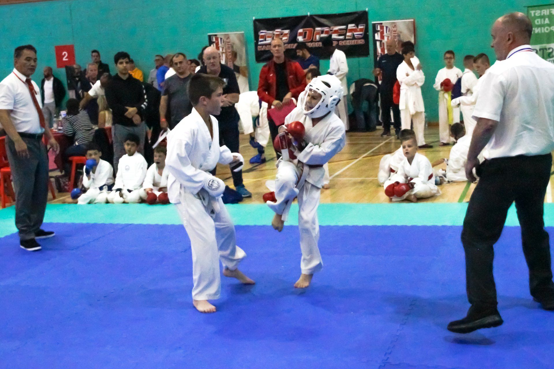 Martial arts sparring match on a blue mat, with two competitors and a referee watching.