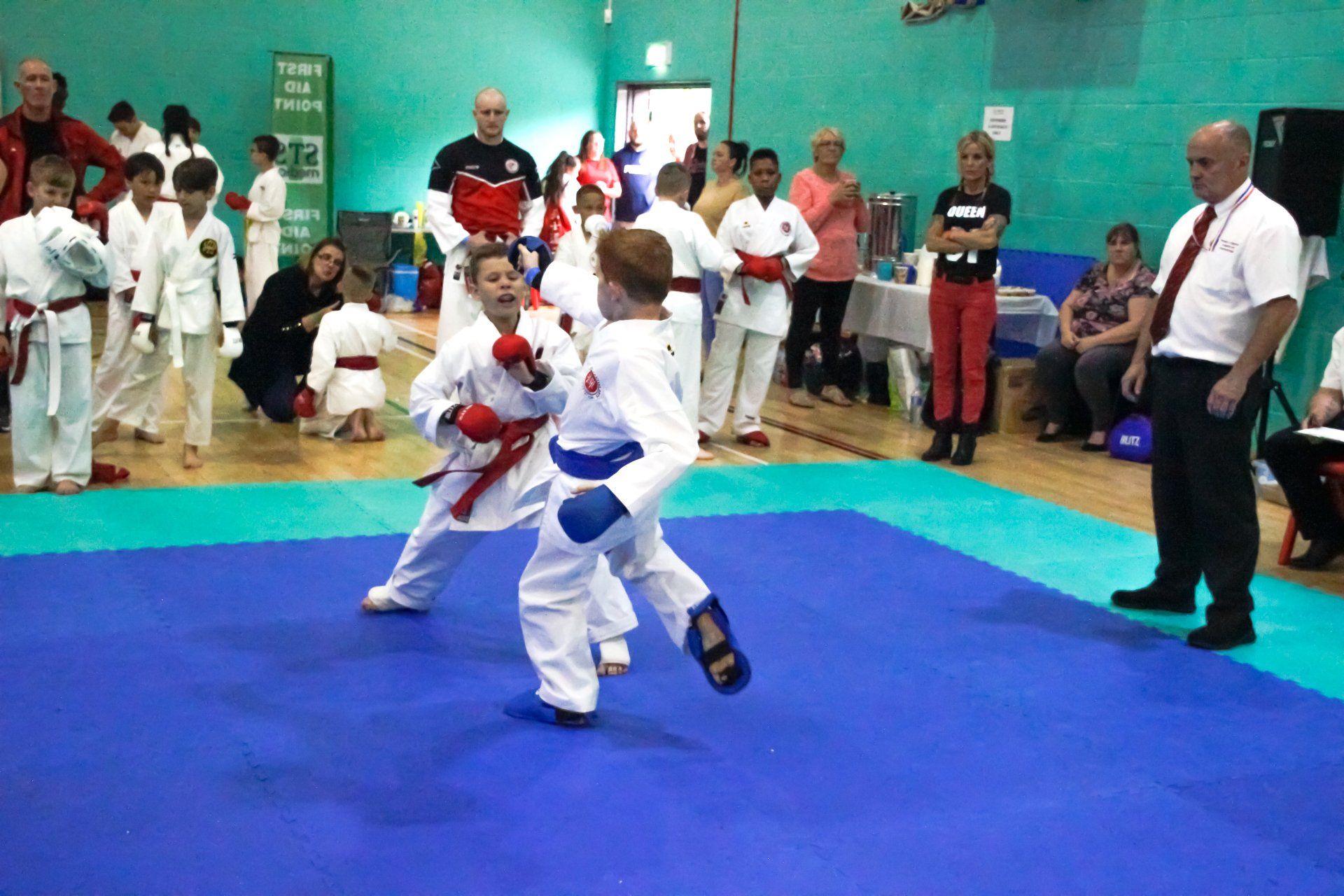 Two karate competitors spar on a blue mat while a referee watches in a gym.
