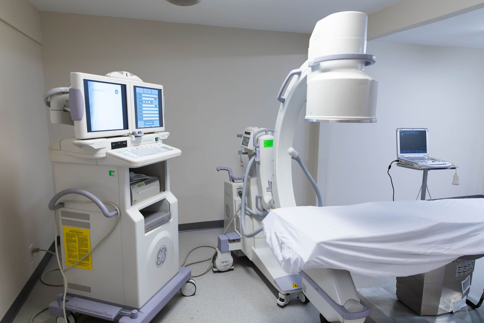 Medical imaging room with a CT scanner, monitor carts, and an examination table covered in white sheets.