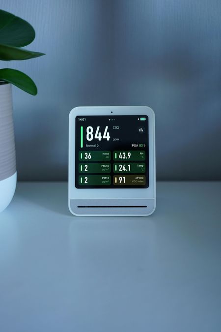White air quality monitor displaying numbers on a table, with a plant to the left.