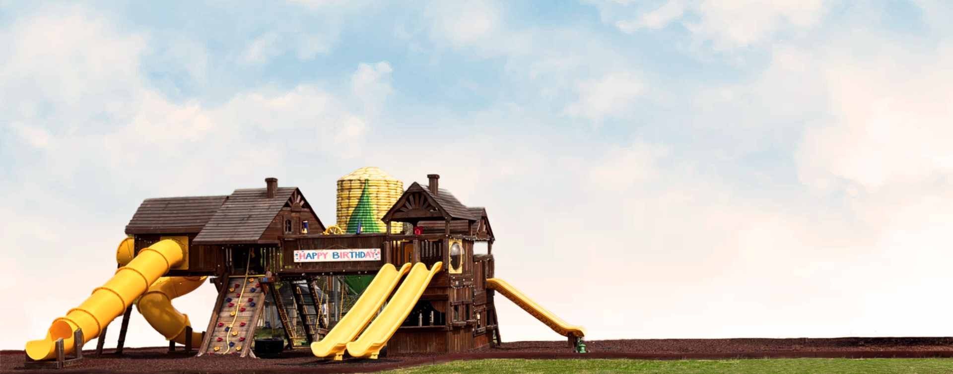 Children's playground with yellow slides & wooden towers on a grassy field under a cloudy sky Grasshopper Farms Knightdale NC