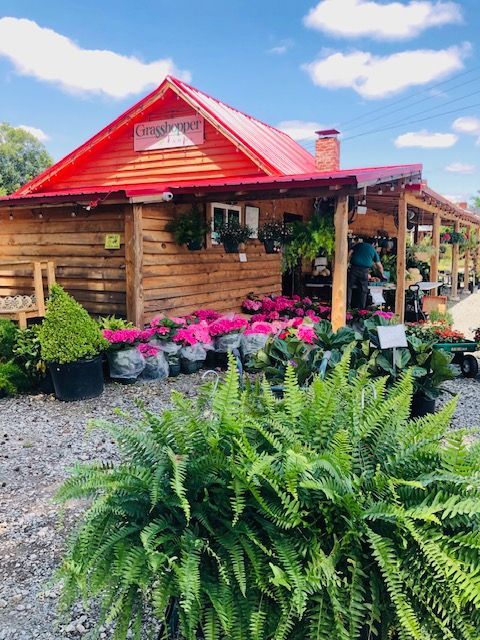 Rustic wooden cabin with bright red roof, surrounded by pink flowers & lush greenery. Grasshopper Farms Knightdale, NC