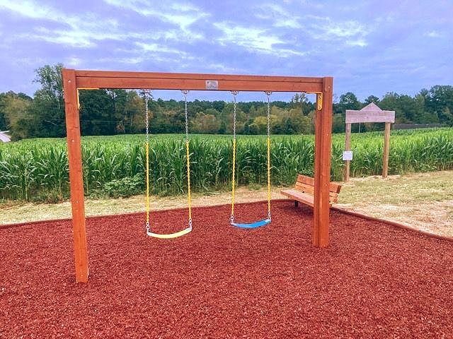 Wooden playground swing set with two seats on red mulch beside a cornfield. Grasshopper Farms Knightdale, NC