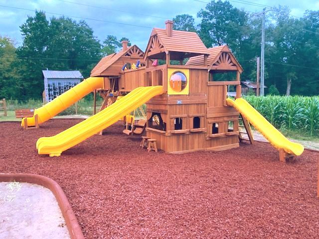 Wooden playground castle with three bright yellow slides on a red mulch surface. Grasshopper Farms Knightdale, NC
