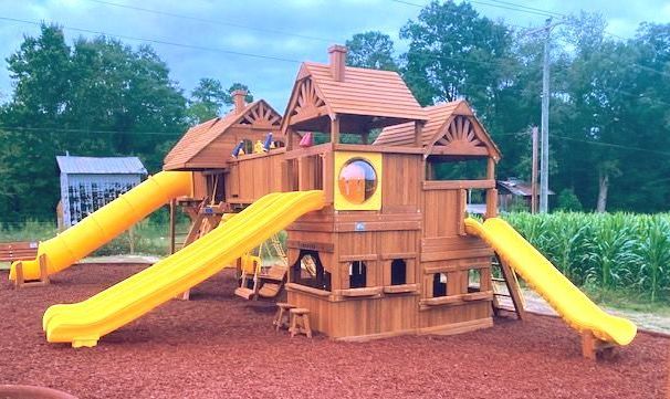Wooden playground playhouse with bright yellow slides on a mulch surface, surrounded by trees and grass. Grasshopper Farms NC