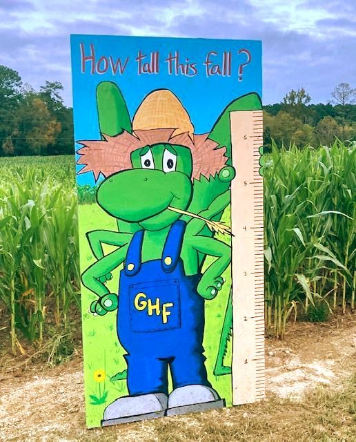 Cartoon green dinosaur in blue overalls stands by a height chart, asking “How tall is tall?” Grasshopper Farms Knightdale, NC