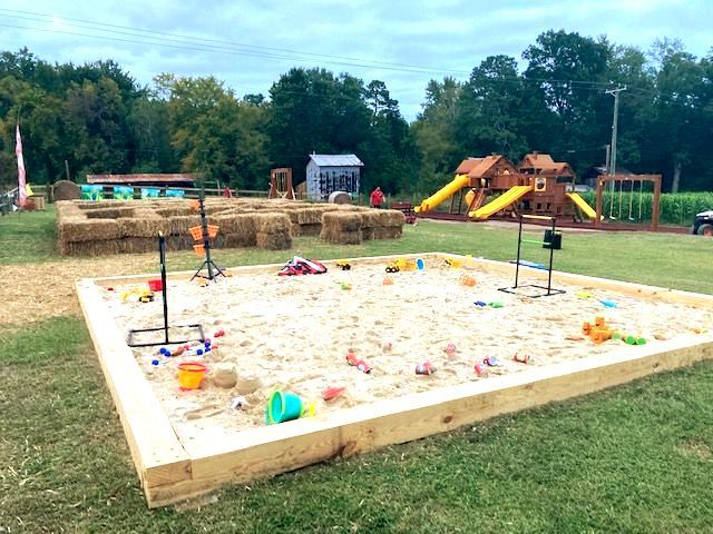 Outdoor sand play area with toys, hay bales, and a playground in the background. Grasshopper Farms Knightdale, NC