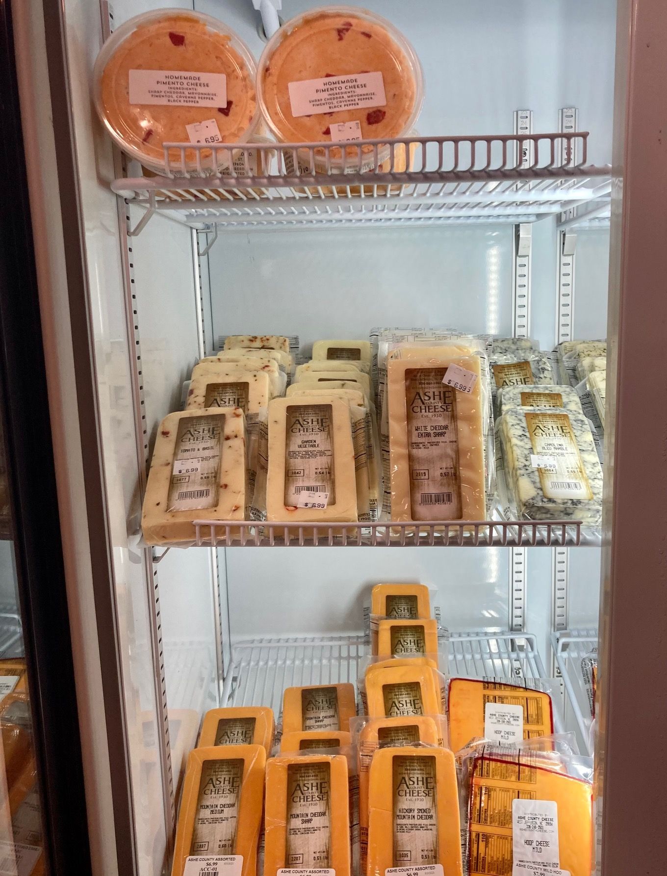 Shelved cheeses in a glass-door display case, with round wheels on top & wrapped blocks below Grasshopper Farms Knightdale NC