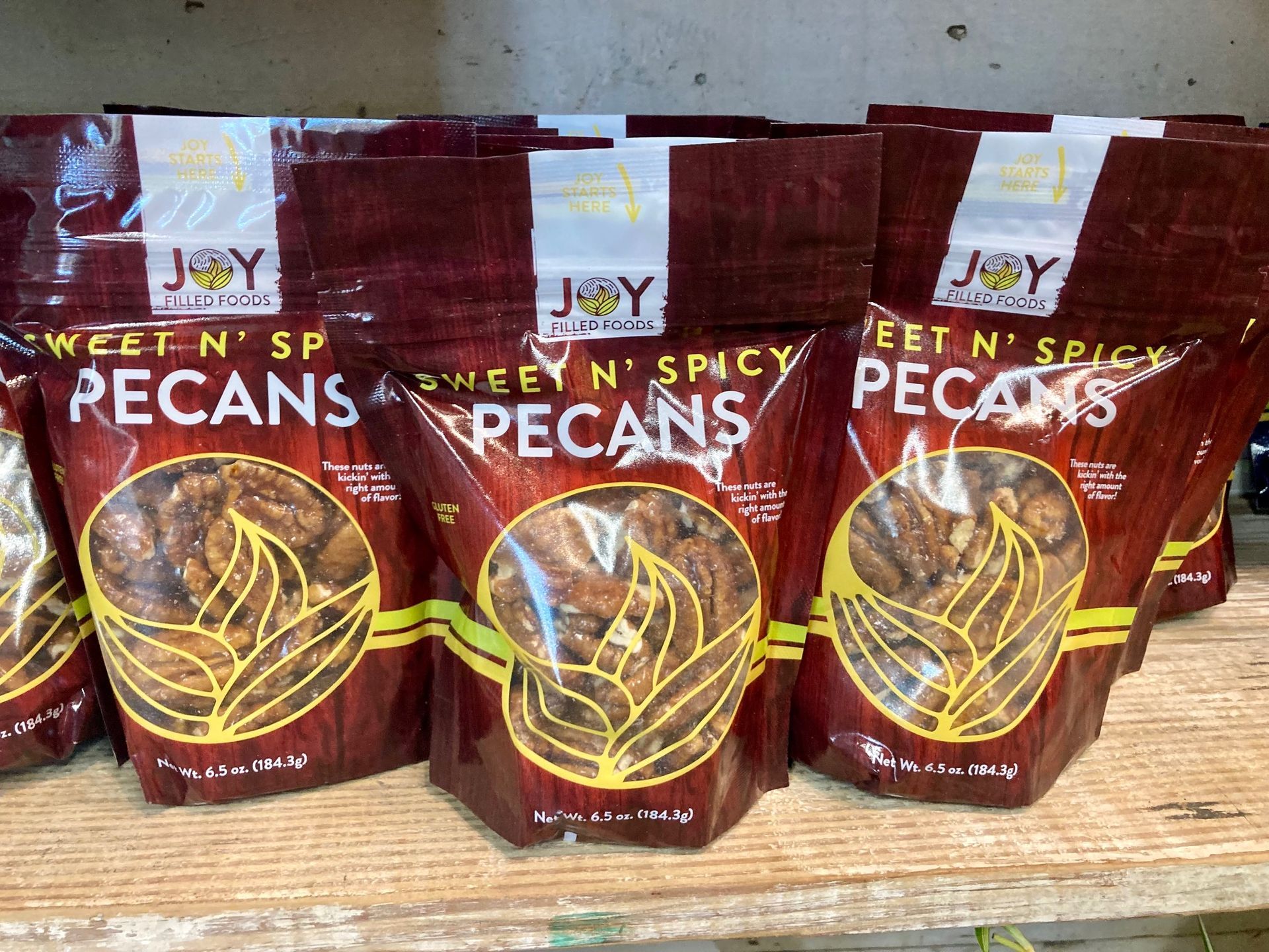 Three bags of Sweet ’n Spicy pecans on a shelf, maroon packaging with gold accents. Grasshopper Farms Knightdale, NC