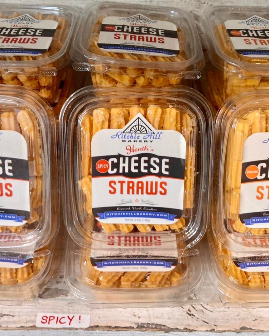 Clear plastic tubs of orange cheese straws on a shelf, labeled “Cheese Straws” & “Spicy” Grasshopper Farms Knightdale NC