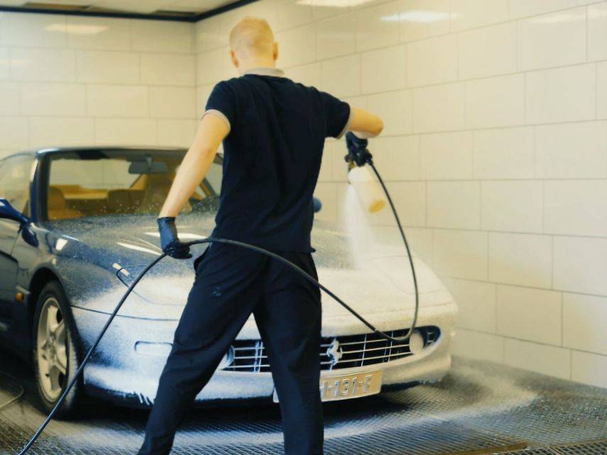 Een persoon wast een zilverkleurige auto met een slang in een betegelde garage bij een autowasstraat.