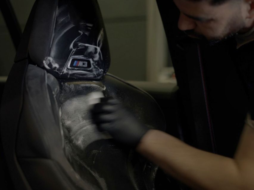 Een persoon maakt met een borstel en een doek een autostoel schoon in het interieur van een voertuig.