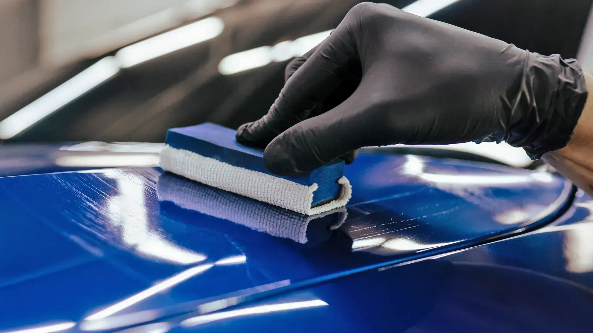 Met een gehandschoende hand poetst hij een glanzende blauwe motorkap met een sponsapplicator.