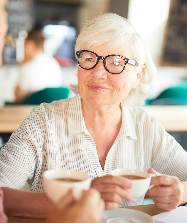 A woman wearing glasses is sitting at a table holding two cups of coffee.