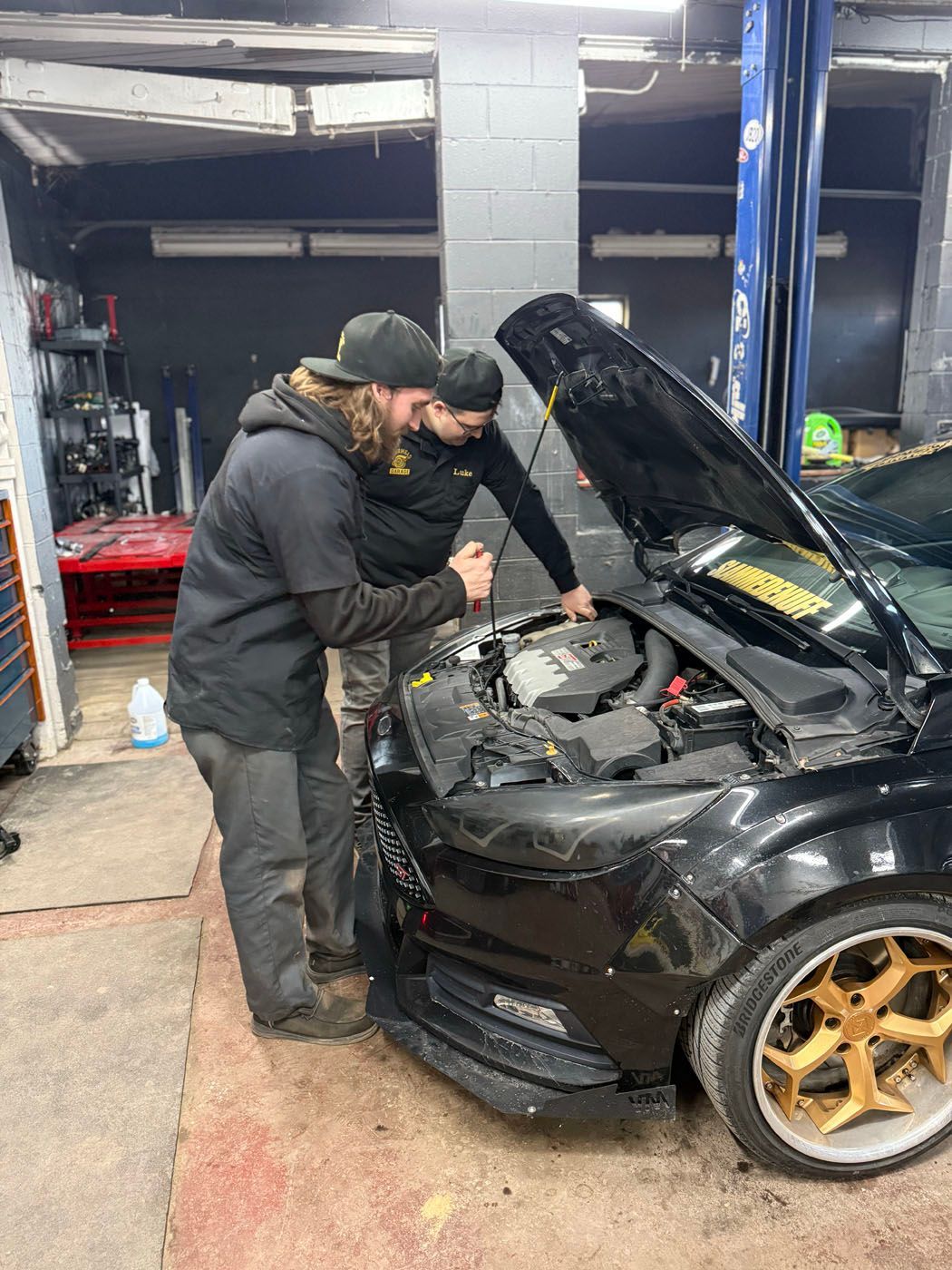 Two mechanics working under the hood of a black car in a garage, with the hood open and gold rims visible