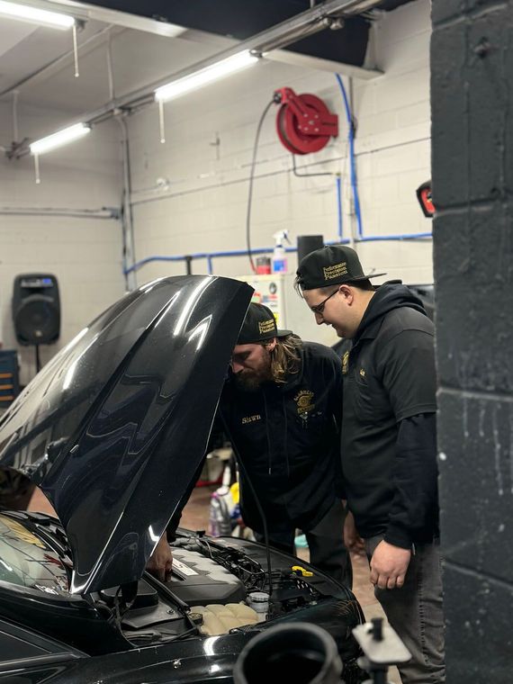 Two people inspecting a car engine in a garage with the hood open.