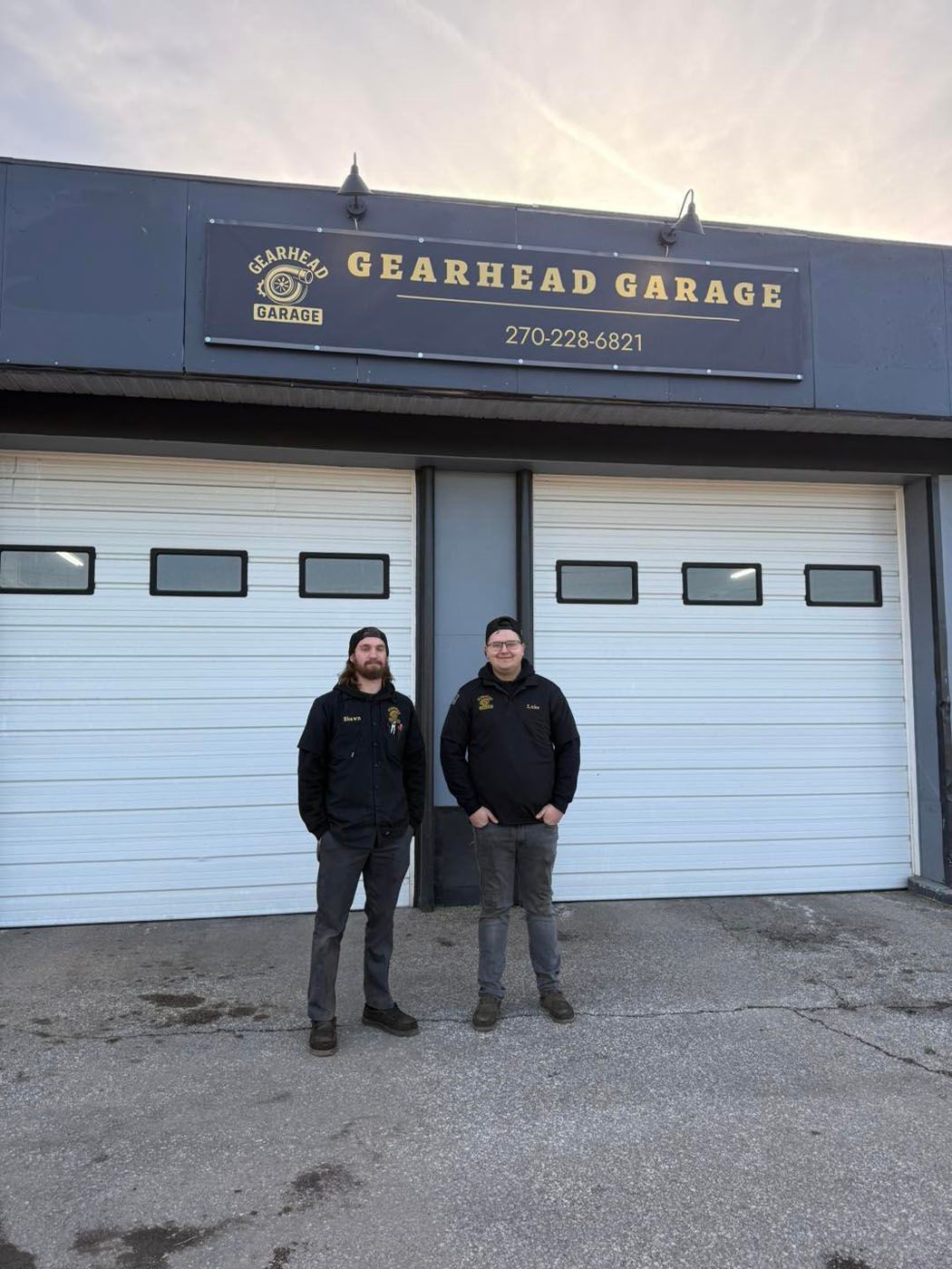 Two people standing outside a garage with blue doors and a “Gearhead Garage” sign.
