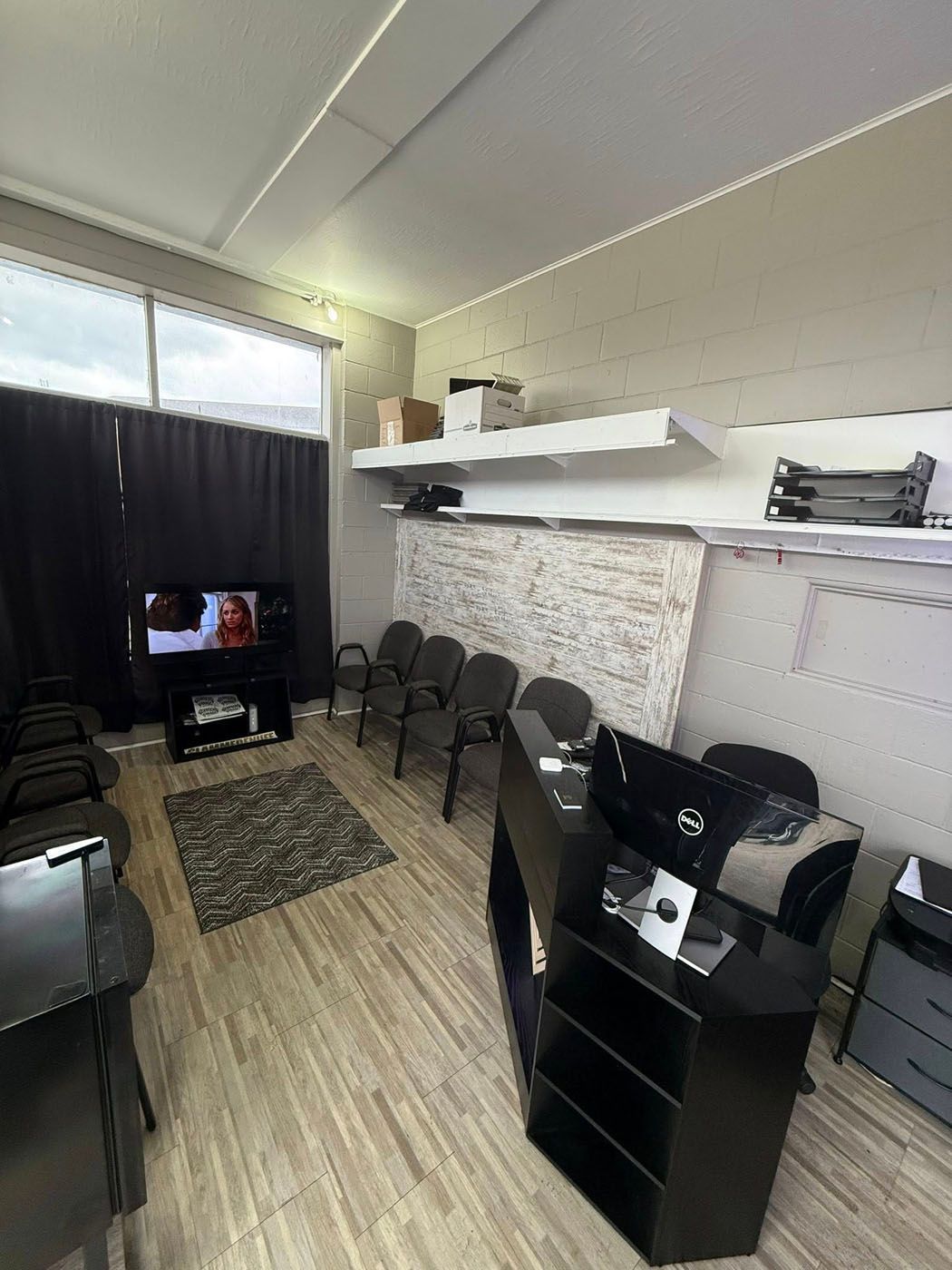 Small waiting room with black chairs, a TV, tiled floor, and shelves along the wall.