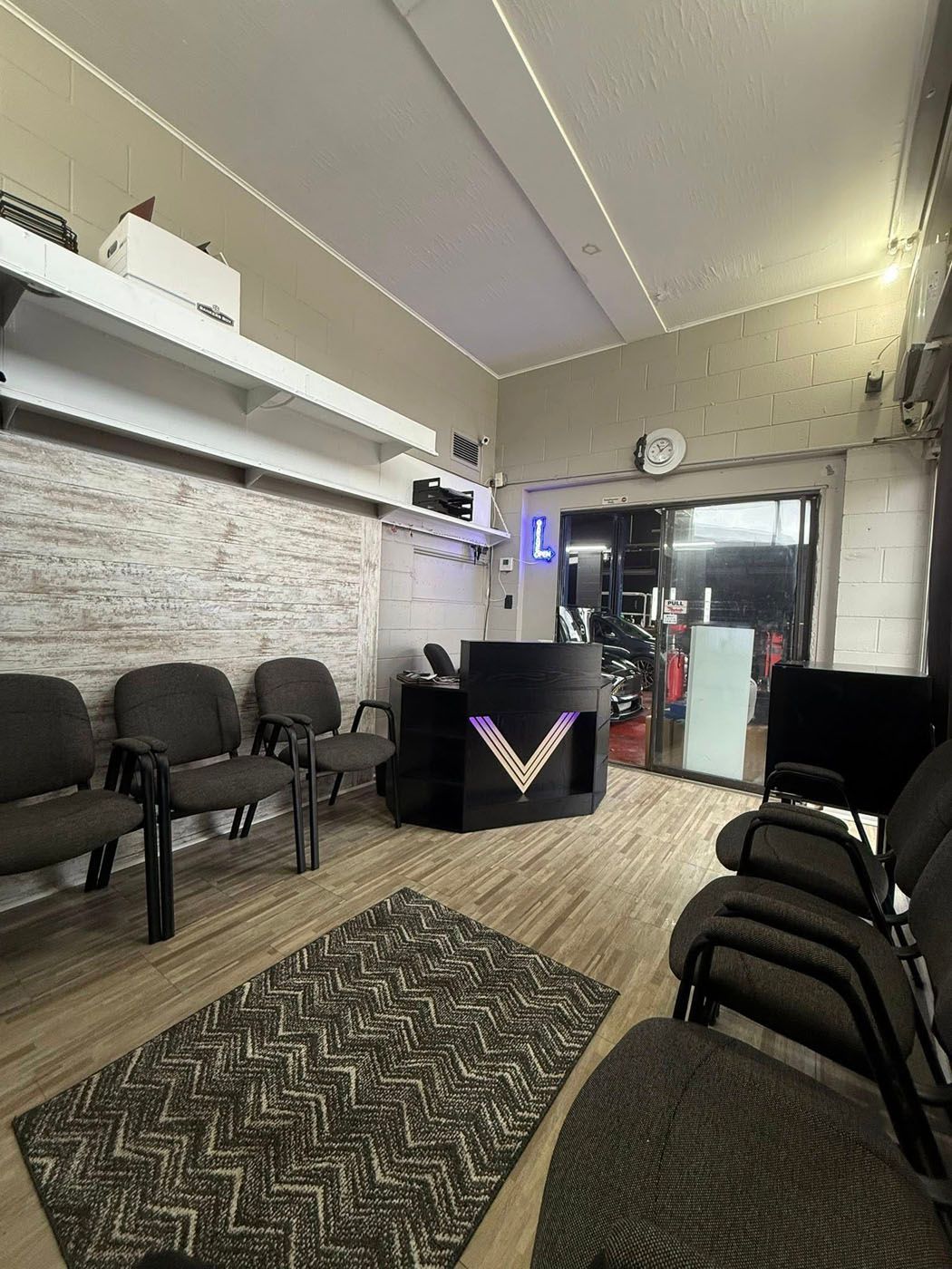 Small waiting room with chairs, patterned rug, reception desk, wall clock, and glass entrance.