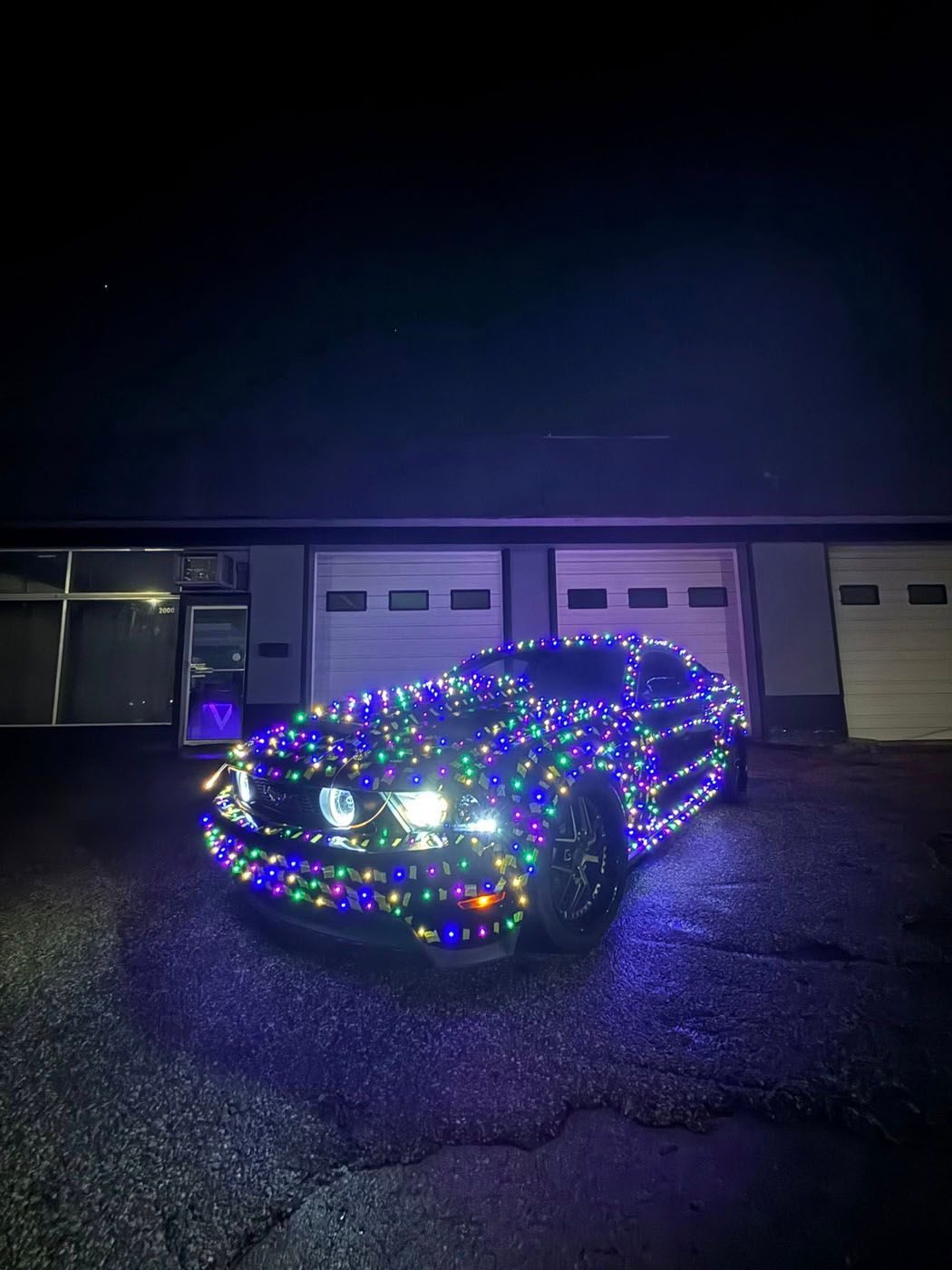 Car covered in colorful holiday lights parked in front of garage doors at night
