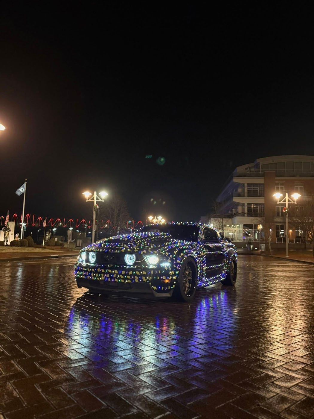 Lit-up black car parked on a wet street at night, reflecting colorful lights.