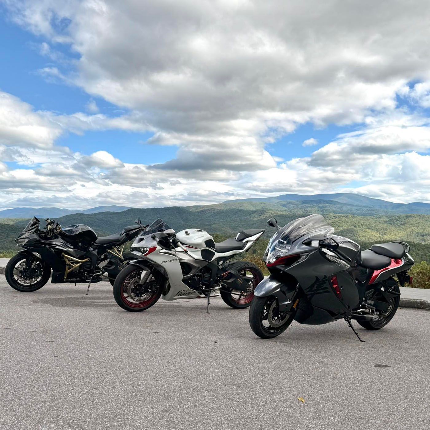 Three sport motorcycles parked on a scenic overlook with mountains and cloudy sky.