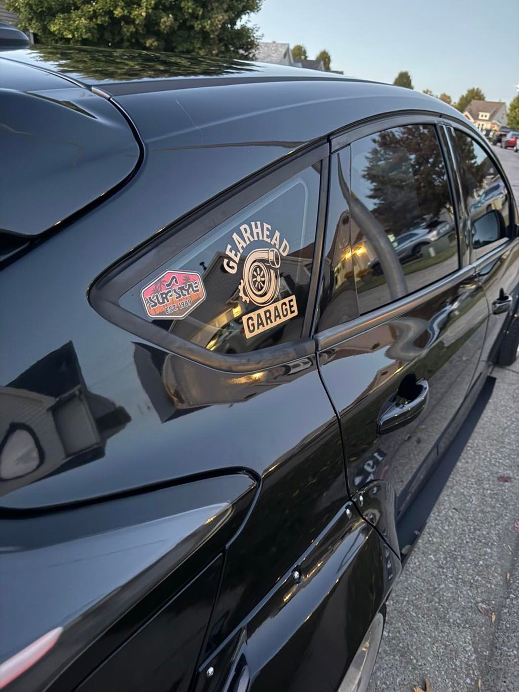 Black car rear window with decals and a gold gear shifter sticker parked on a street.