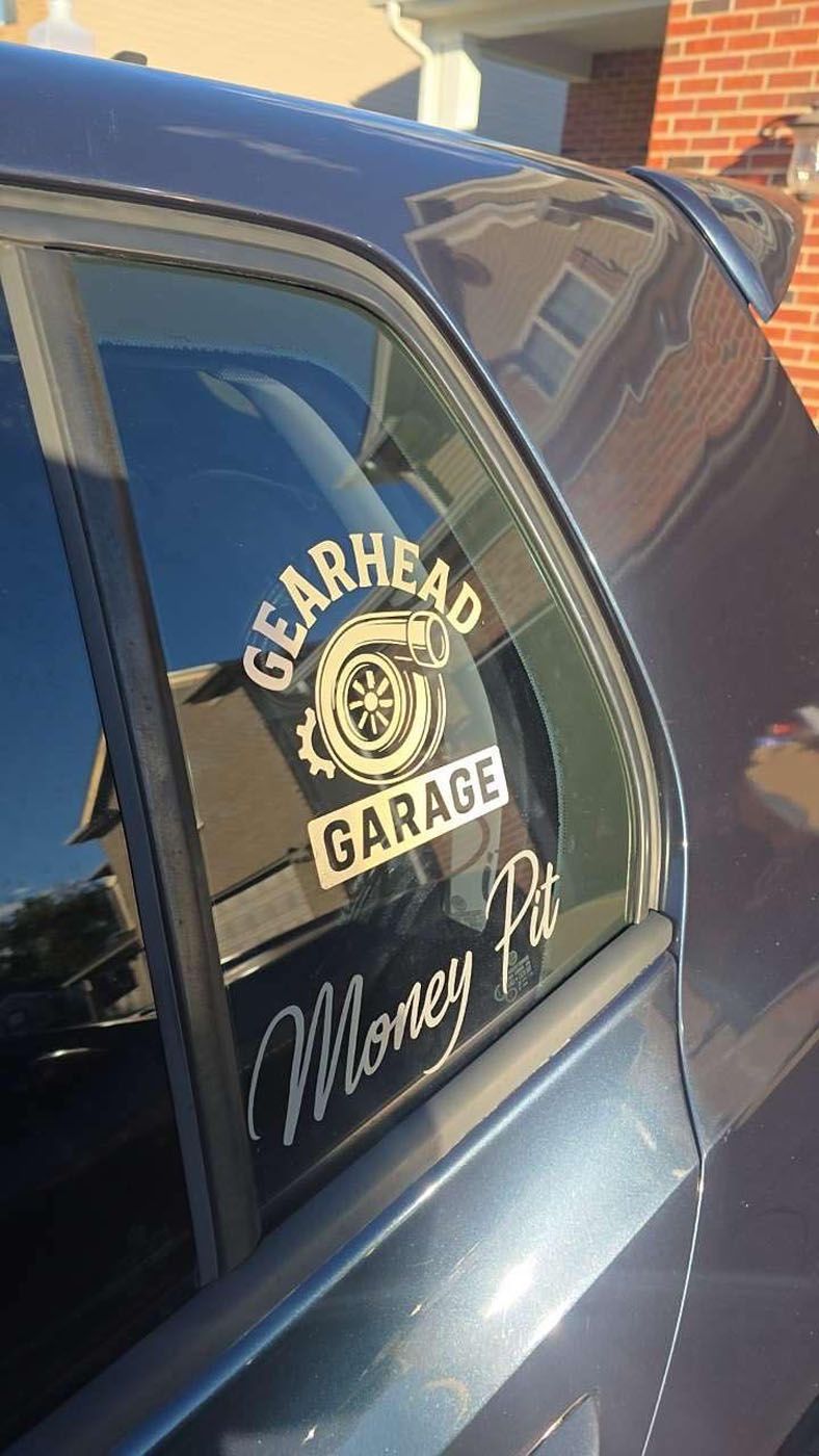 Blue car window with a “Gearhead Garage” Monkey logo decal at a brick building.