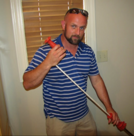 Man holding a red-handled broom or mop, standing indoors in a blue striped polo and khaki pants.