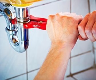 Hands using a red pipe wrench to tighten a chrome sink drain pipe under a sink.