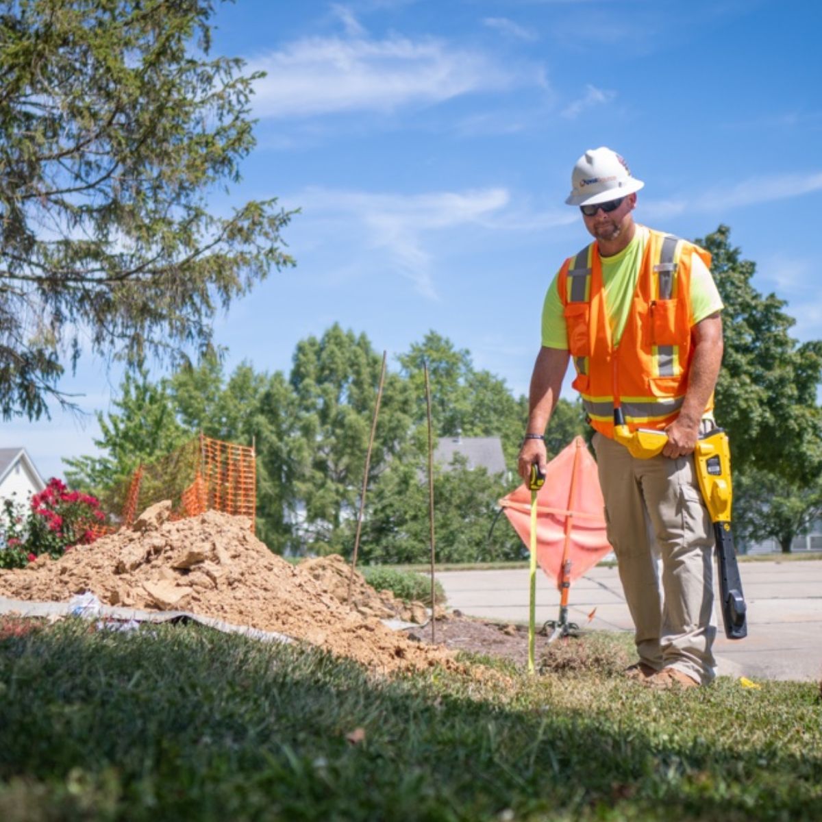 A man in an orange vest and hard hat is standing in the grass holding a yellow device.