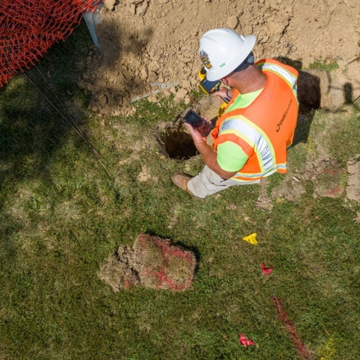 A construction worker is sitting in the grass looking at his cell phone.