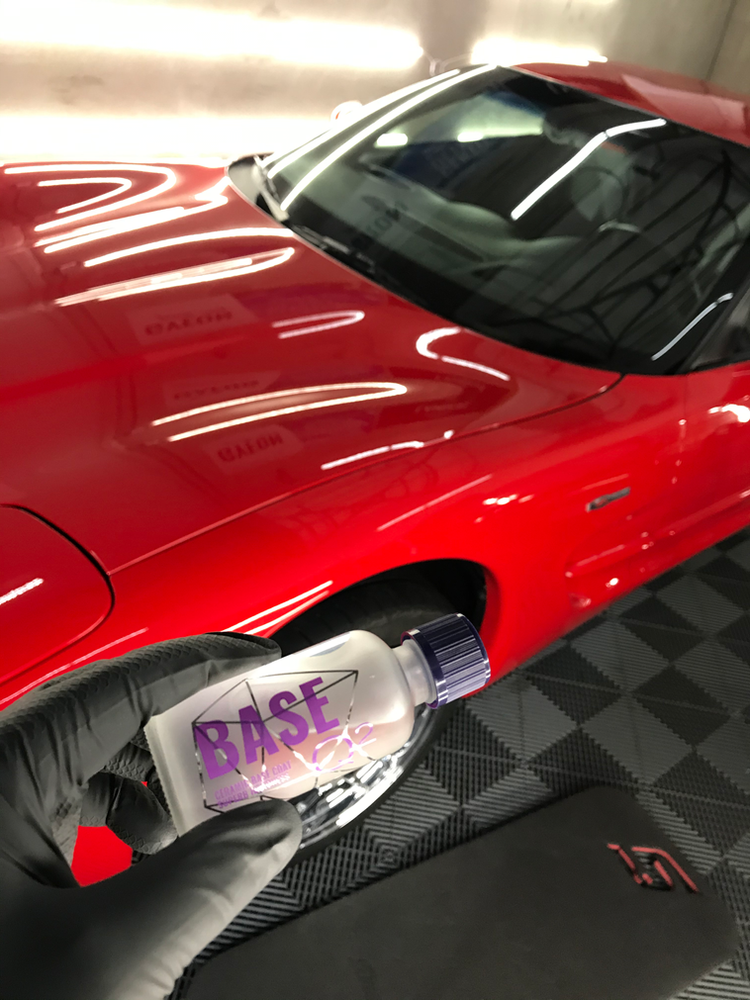 A person is holding a bottle of base in front of a red car.