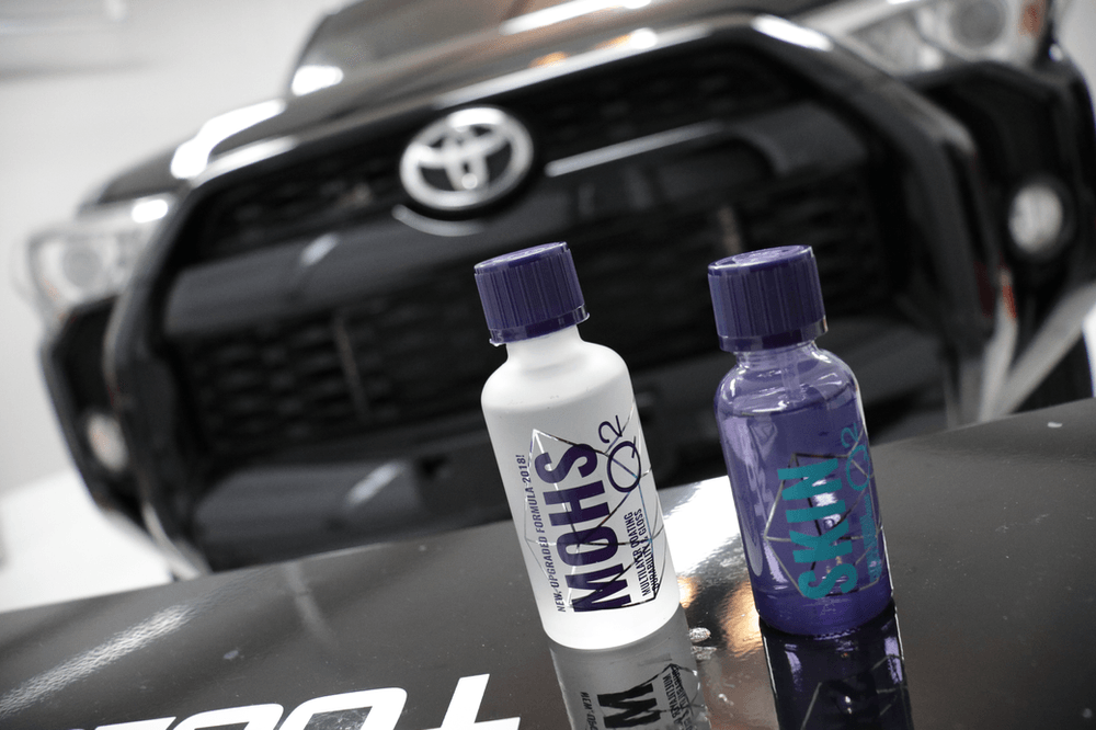 Two bottles of polish are sitting on a table in front of a car.