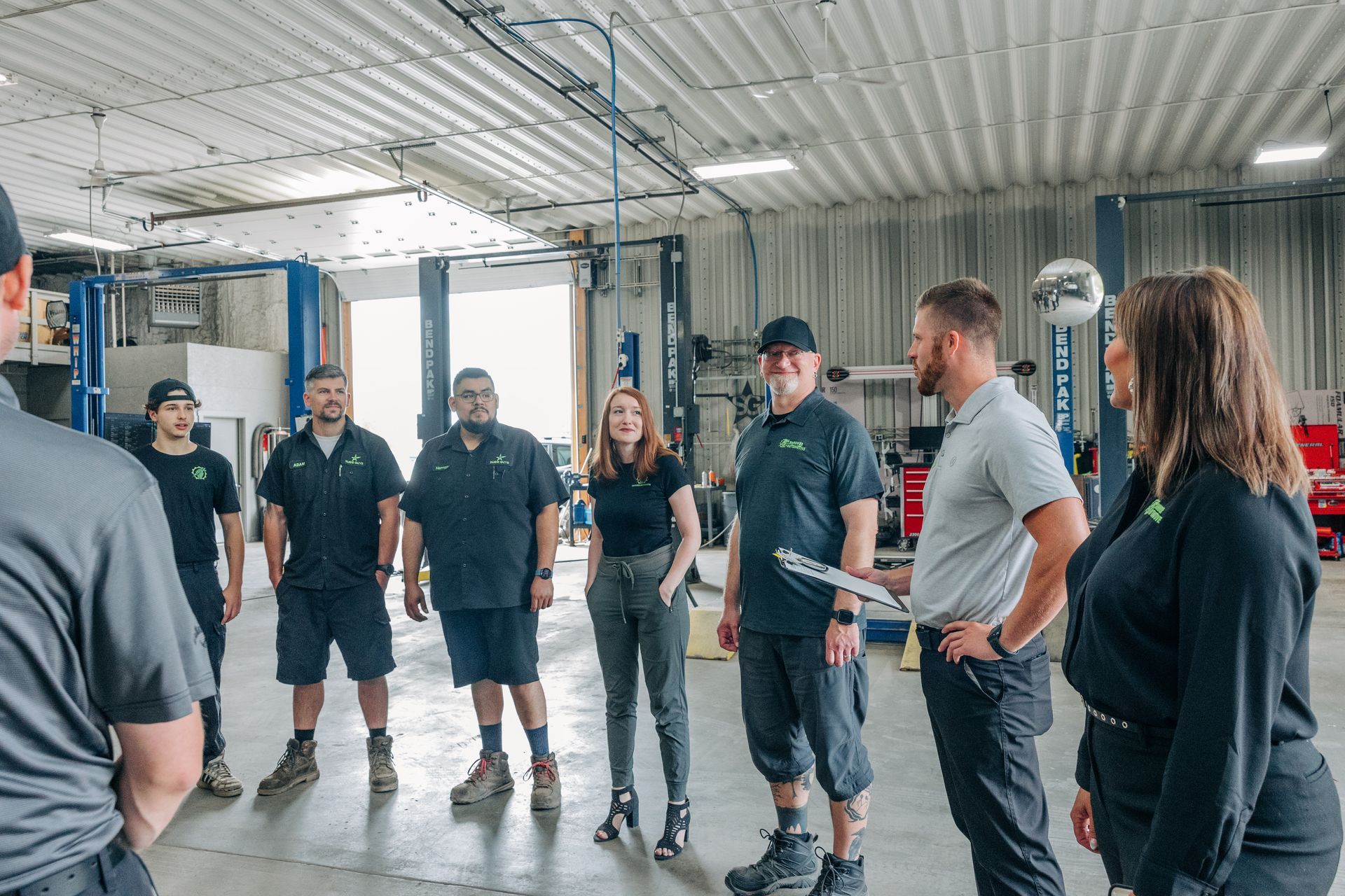 A group of mechanics in a garage stands in a circle, listening to a woman.