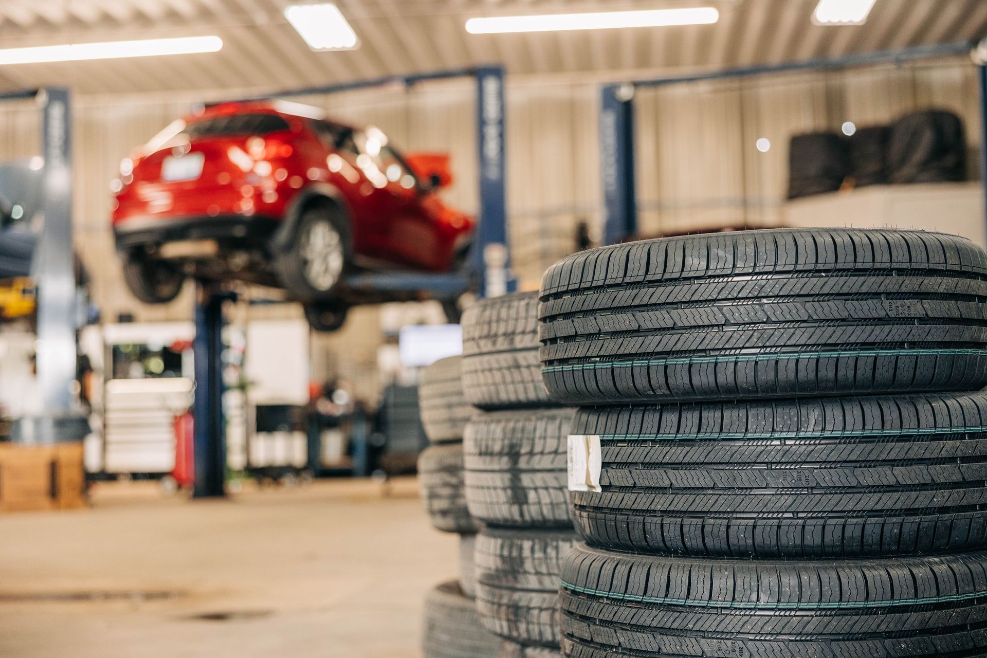 Stack of new tires in foreground, red car elevated on a lift in a repair shop. | Empower Automotive