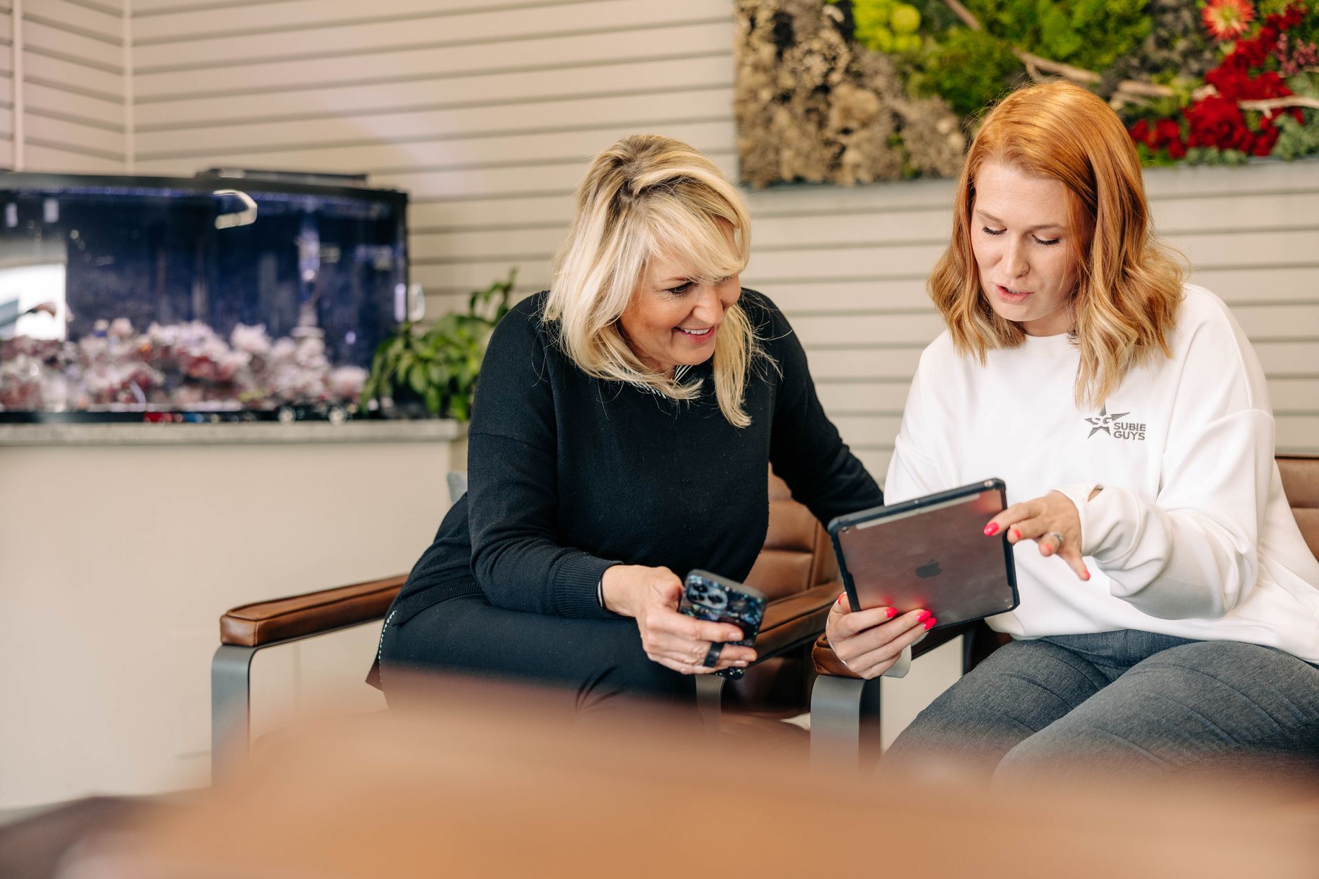 Two women looking at a tablet in an office. One points, both smile. Background: aquarium, plants, neutral walls.