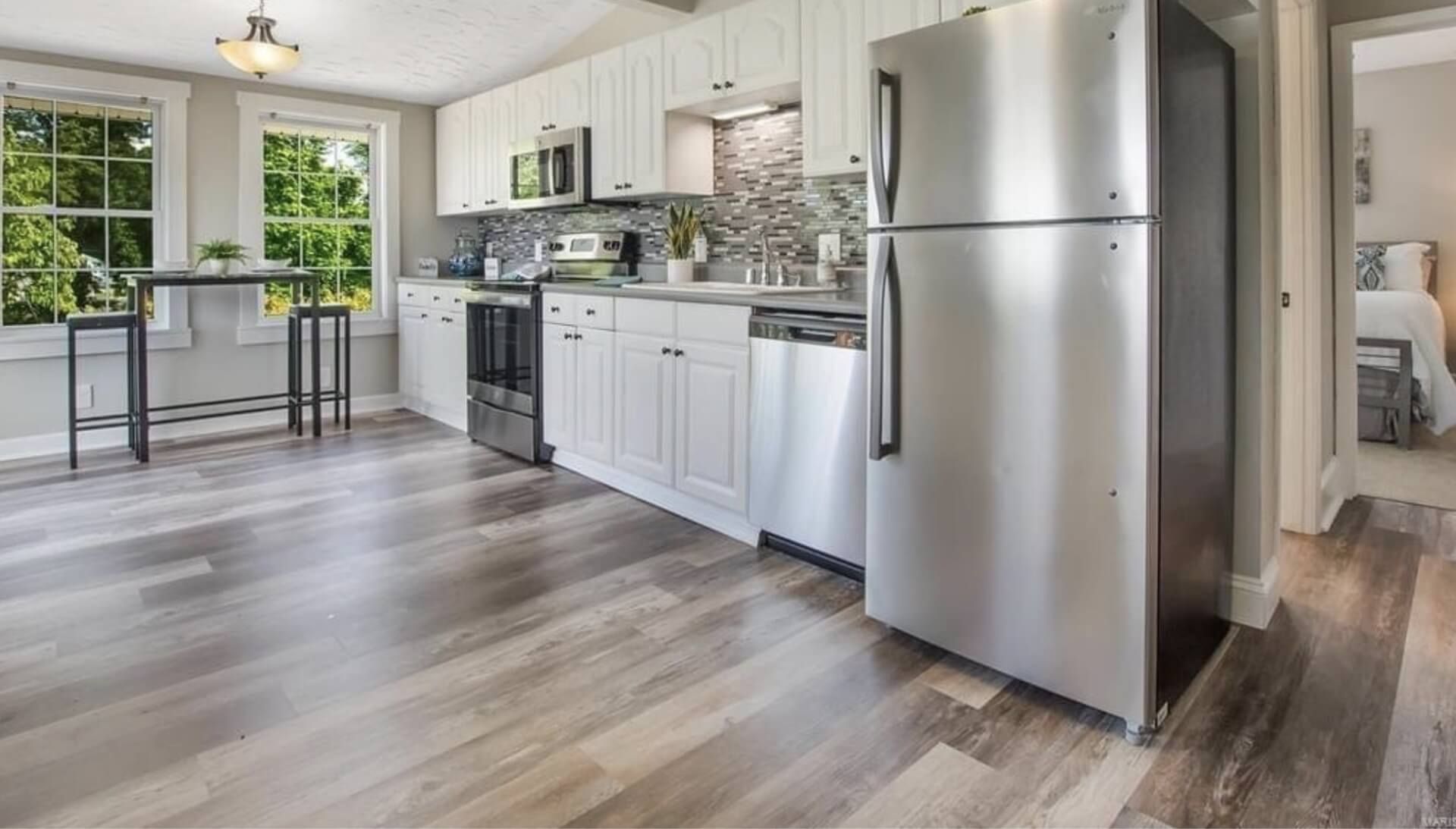 Bright modern kitchen with stainless steel fridge, white cabinets, and wood-look flooring by a window.