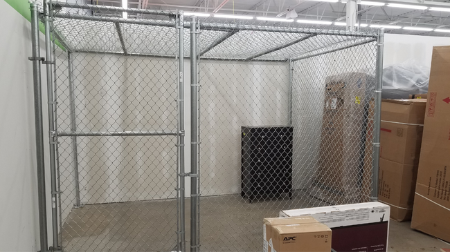 A chain-link cage in a store holds boxes and a black container. The cage has a door.